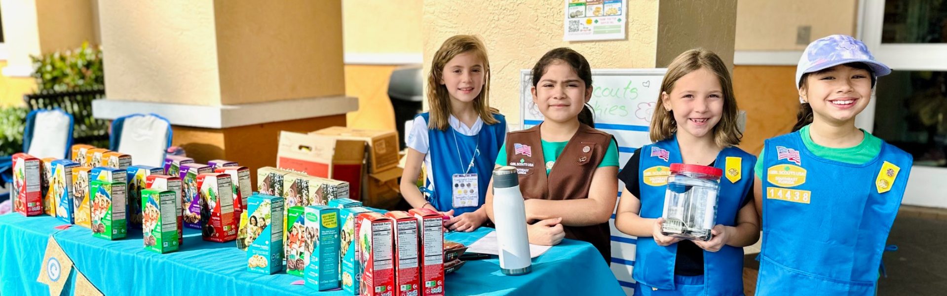  girl scouts selling cookies with one girl in front of booth holding sign that says "girl scout cookie proceeds stay local" 
